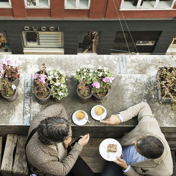 Två personer njuter av kaffe på takterrassen i Köpenhamns Illum med utsikt och blommor i bakgrunden.