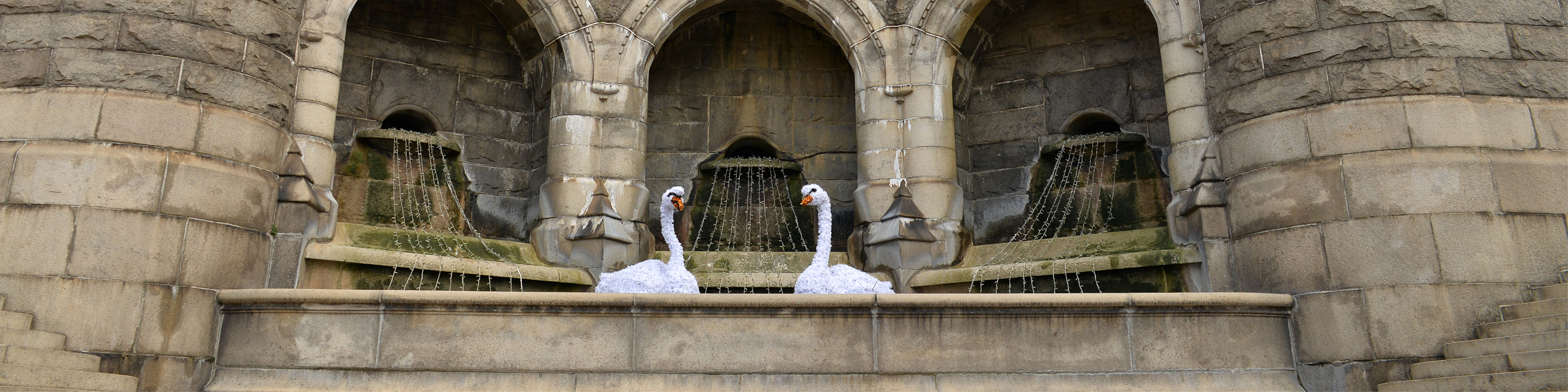 To hvide svanelignende skulpturer foran en gammel stenfontæne med buer og løbende vand i baggrunden.