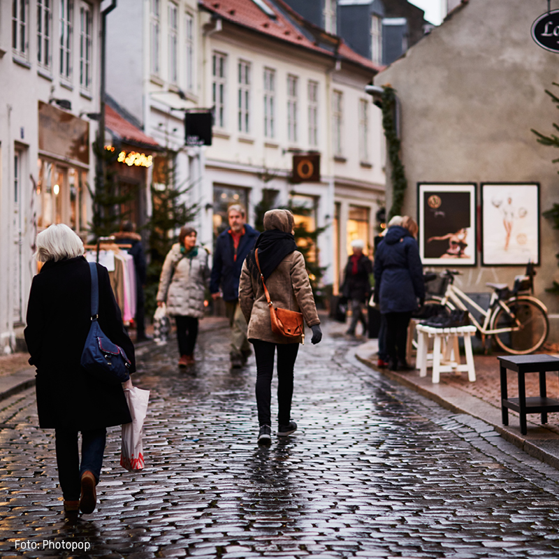 Folk går på brostensbelagt gade i en dansk by, iført vinterjakker og bæreposer, mens de handler i butikker.