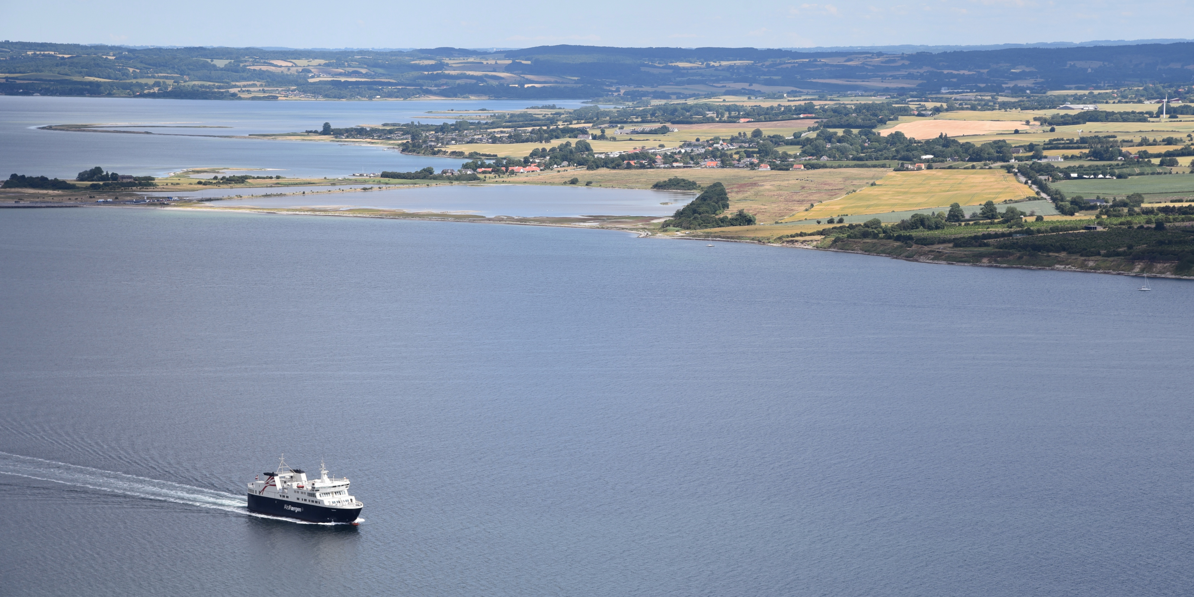 Aarhus færge krydser det åbne farvand set fra luften med lystbådehavn og kystlandskab i baggrunden.