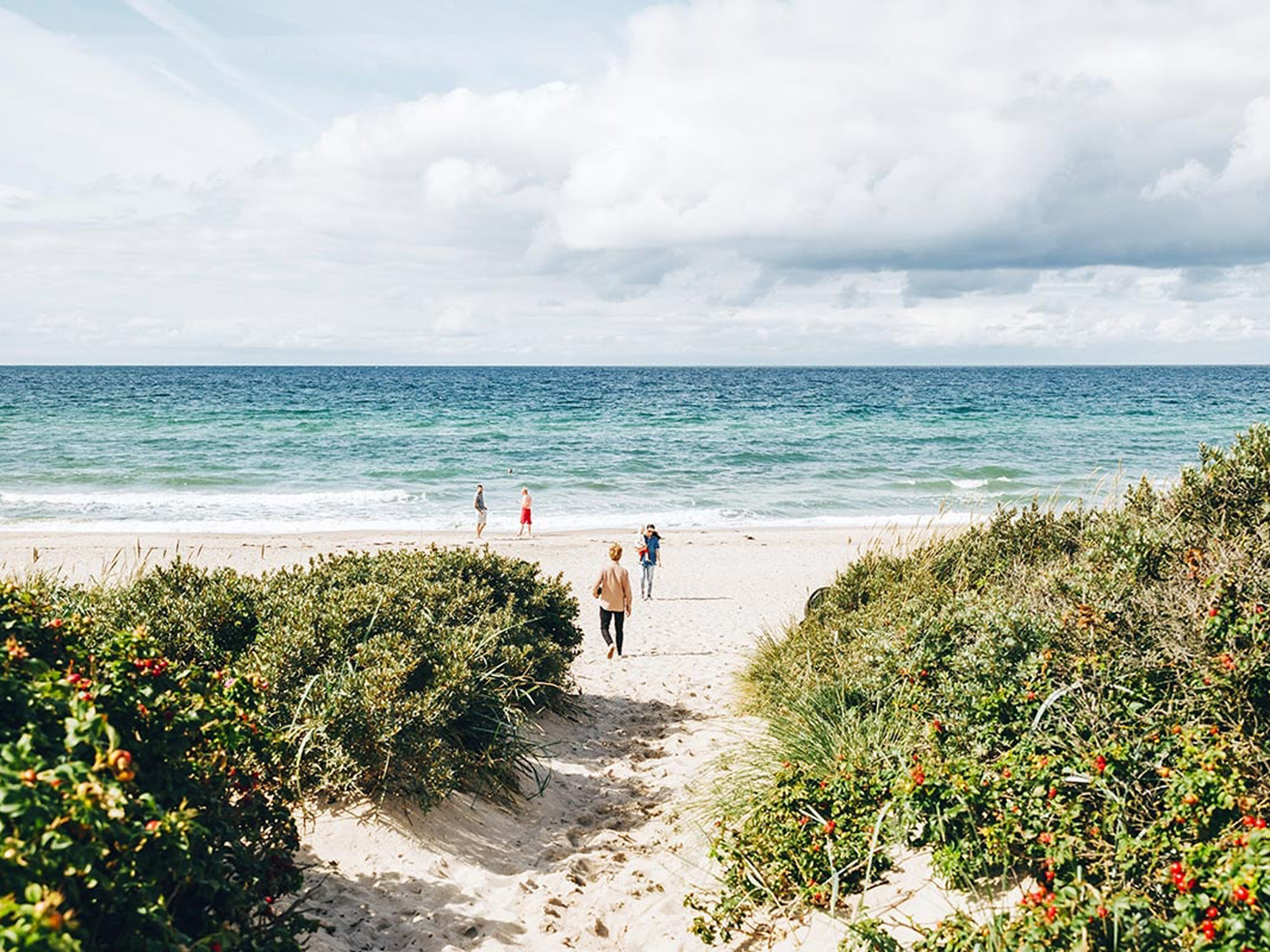 Besökare promenerar ner till en avskild strand vid danska rivieran med lugna vågor och klar himmel i bakgrunden.