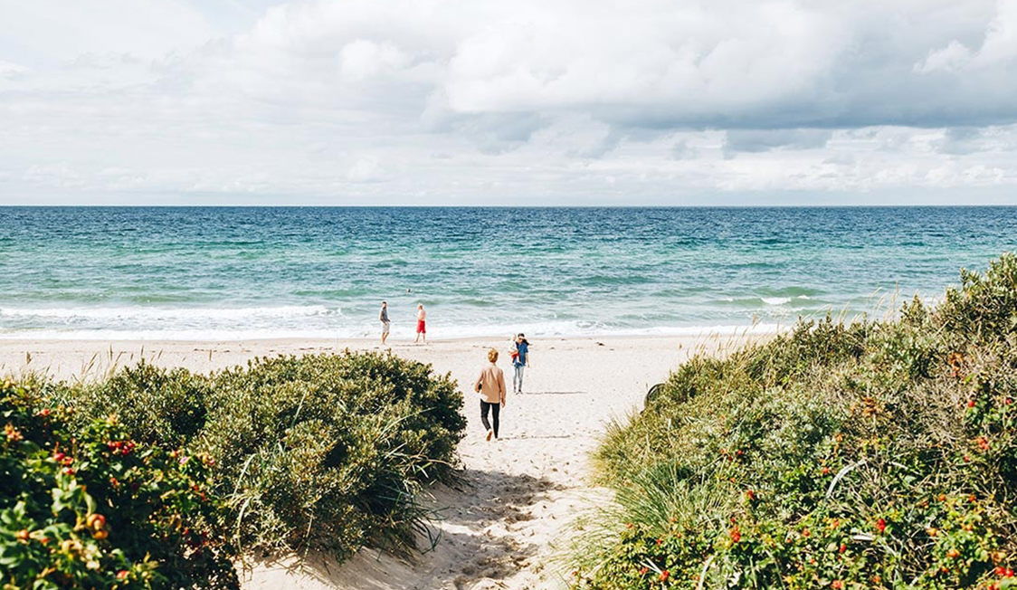 Besökare promenerar ner till en avskild strand vid danska rivieran med lugna vågor och klar himmel i bakgrunden.