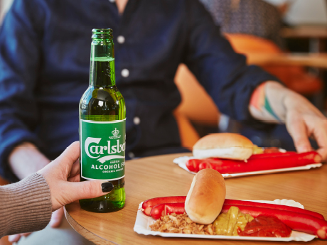 Person håller Carlsberg alkoholfri öl vid bord med två hot dogs i pappersformar.