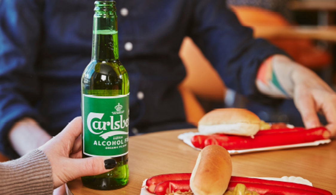 Person håller Carlsberg alkoholfri öl vid bord med två hot dogs i pappersformar.