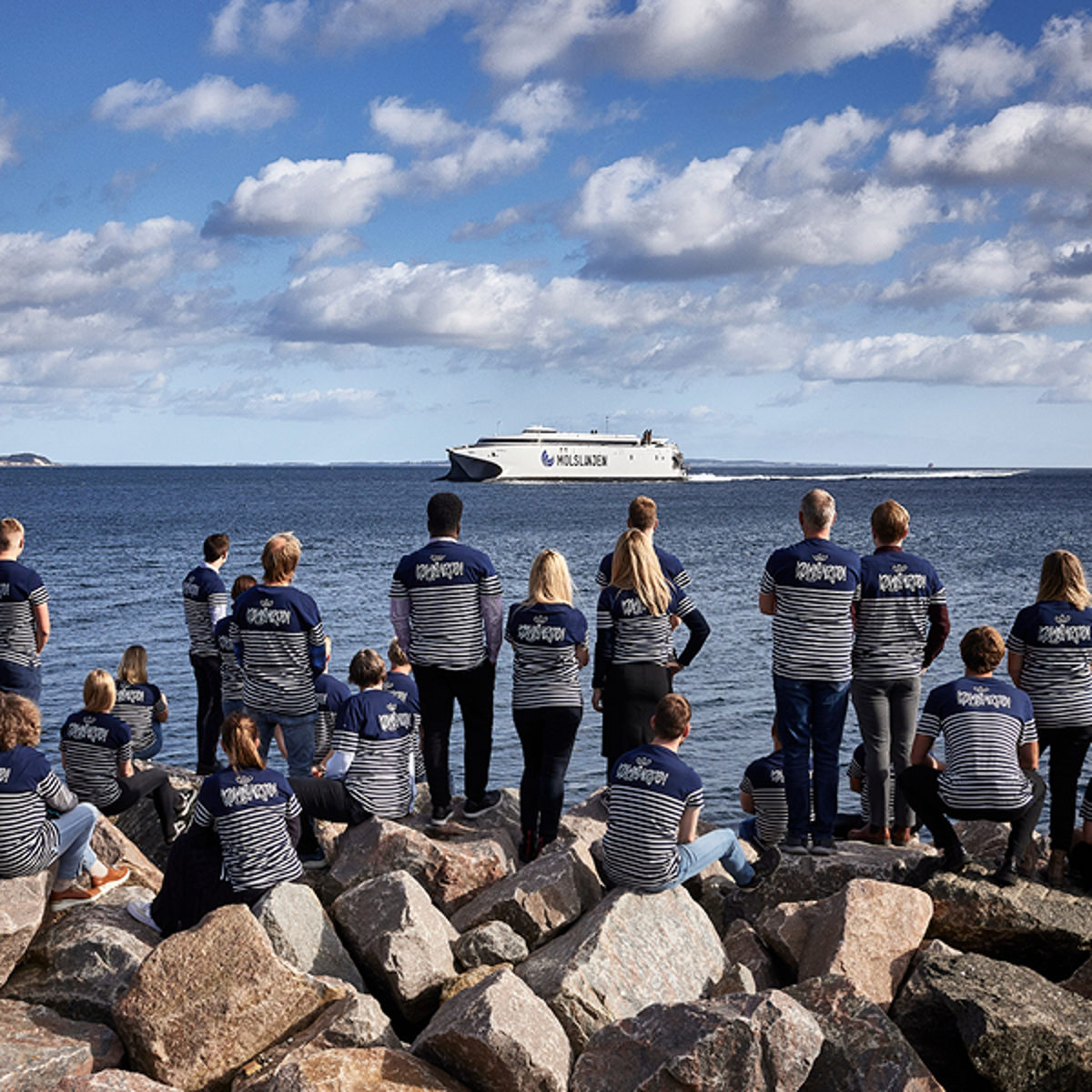 Molslinjen-færge sejler på havet, mens en gruppe mennesker i stribede trøjer ser fra kysten.