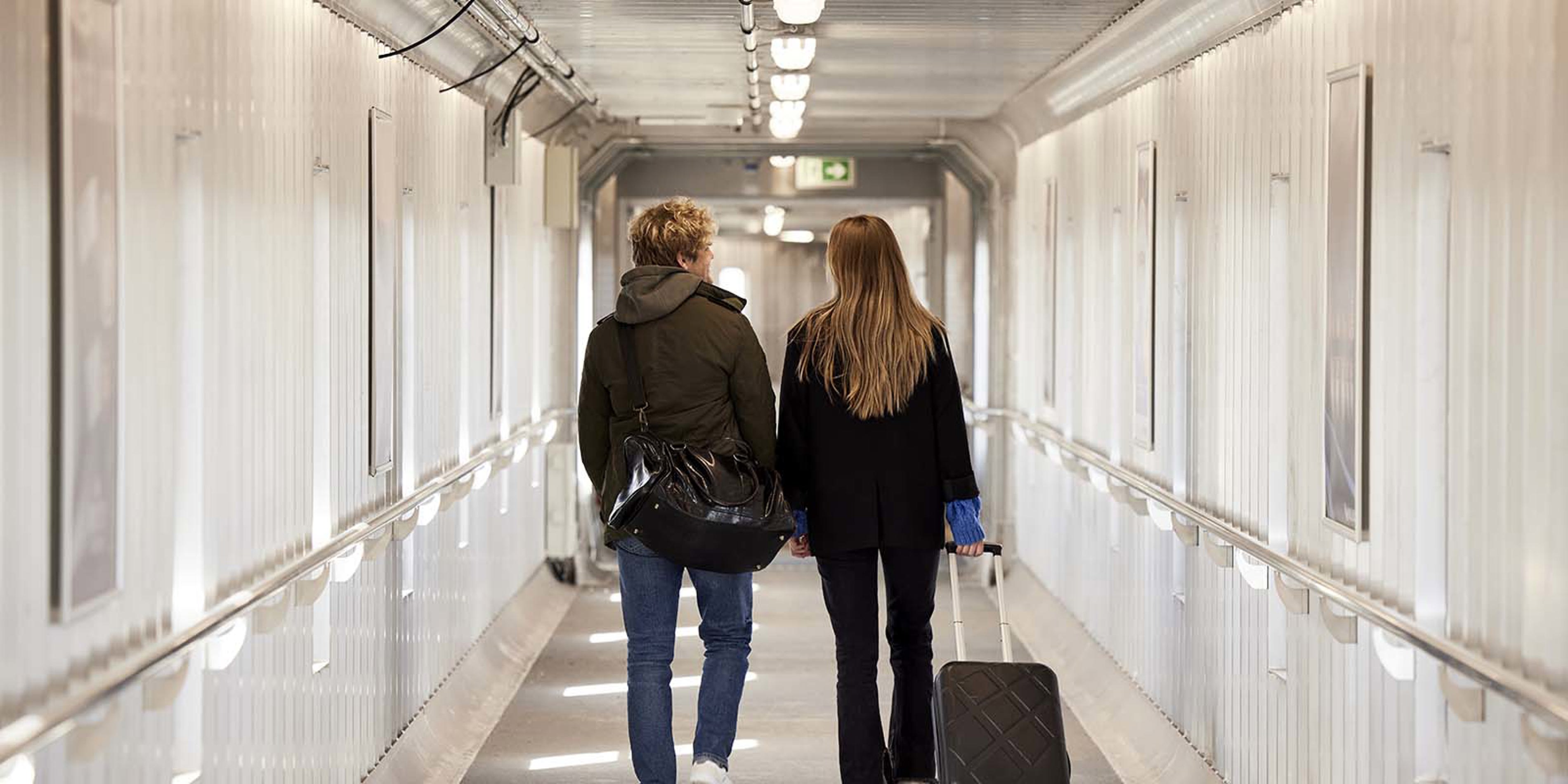 To personer går gennem en lys gang med bagage i hånd, set bagfra.