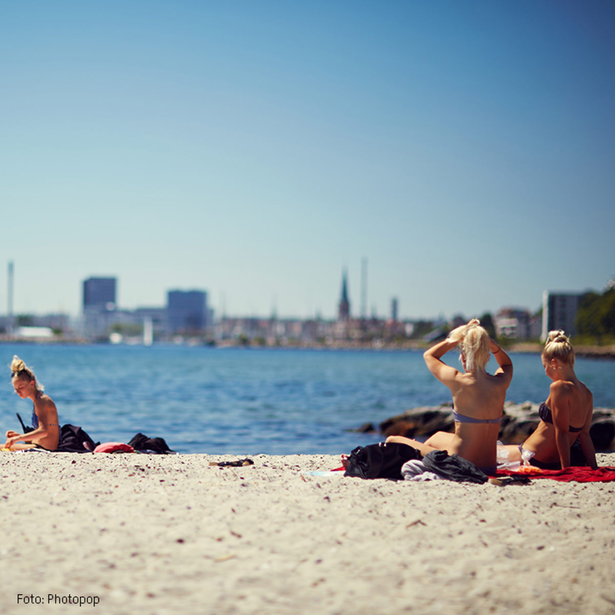 Kvinder solbader på Hellerup Strand med udsigt til København i baggrunden på en solrig sommerdag.