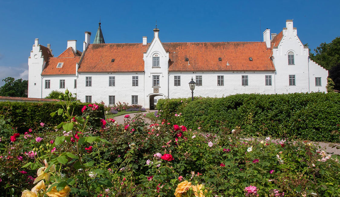 Renaissance-jagtpalæ med rødt tegltag og hvidkalkede vægge set over en blomstrende rosenhave og blå himmel.