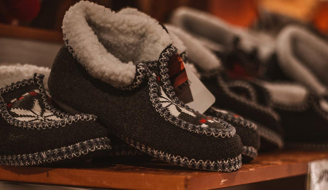 Hjemmestrikkede, grå hjemmesko med hvid foer og traditionelt mønster udstillet på træhylde i butik.