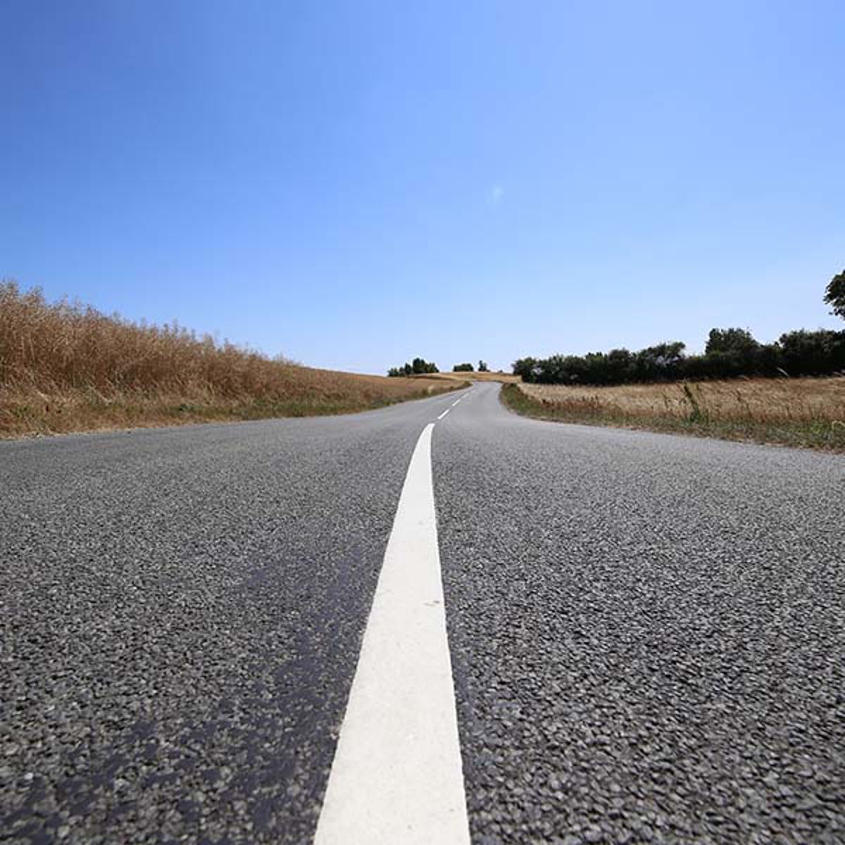 Lang, lige landevej strækker sig gennem landskabet under en klar blå himmel, set fra vejniveau.