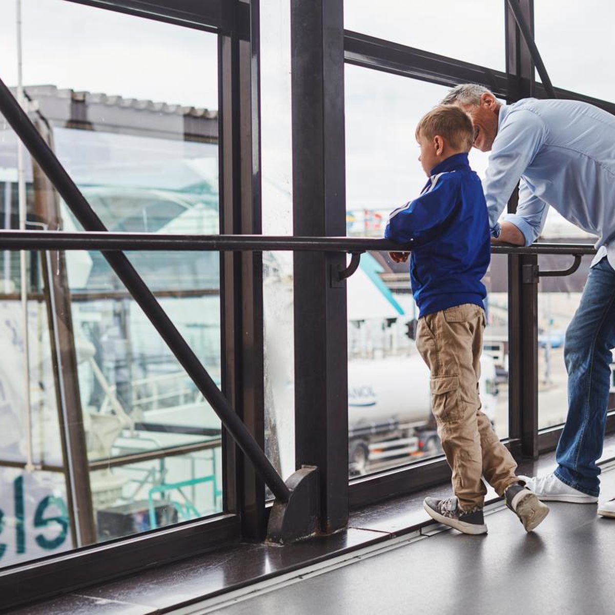 Man and boy in blue jackets look out over the harbor area through a large glass window.