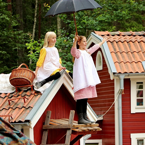 To piger i gammeldags kjoler sidder på hustag med kurv og paraply, omgivet af grønne træer.