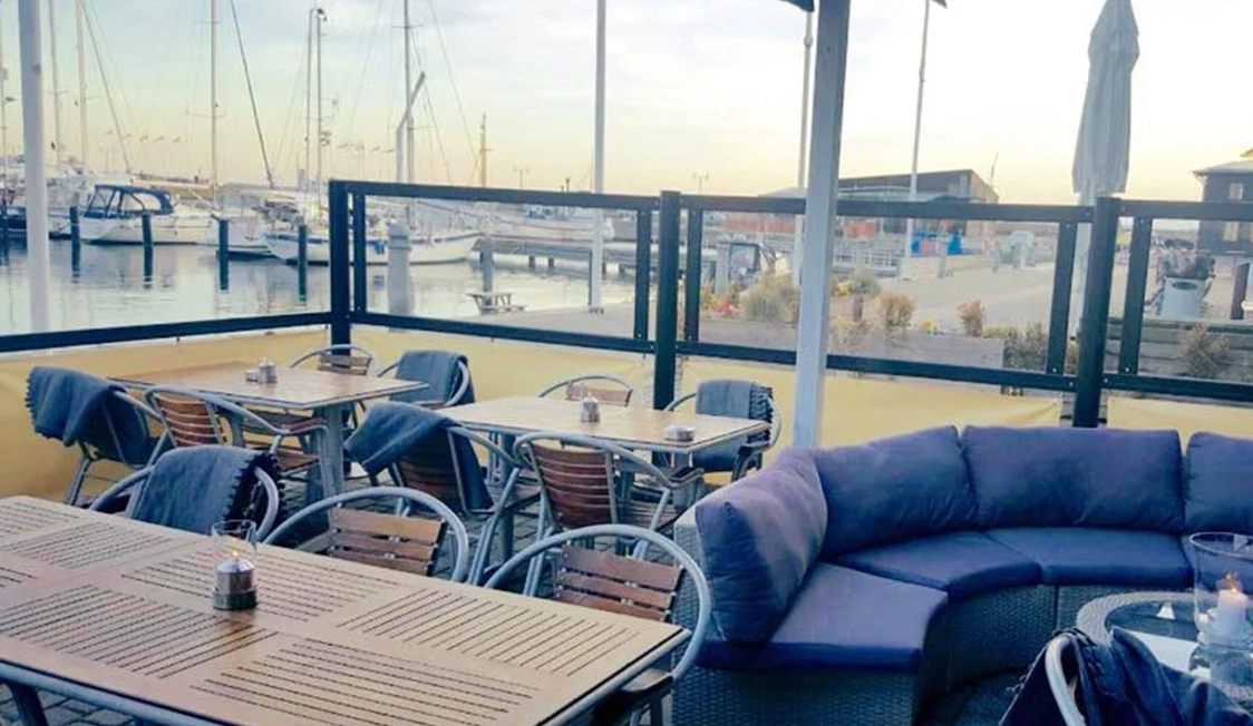 Udendørs terrasse med træborde og stole, udsigt over lystbådehavn, dækket af tæpper og puder til komfort.