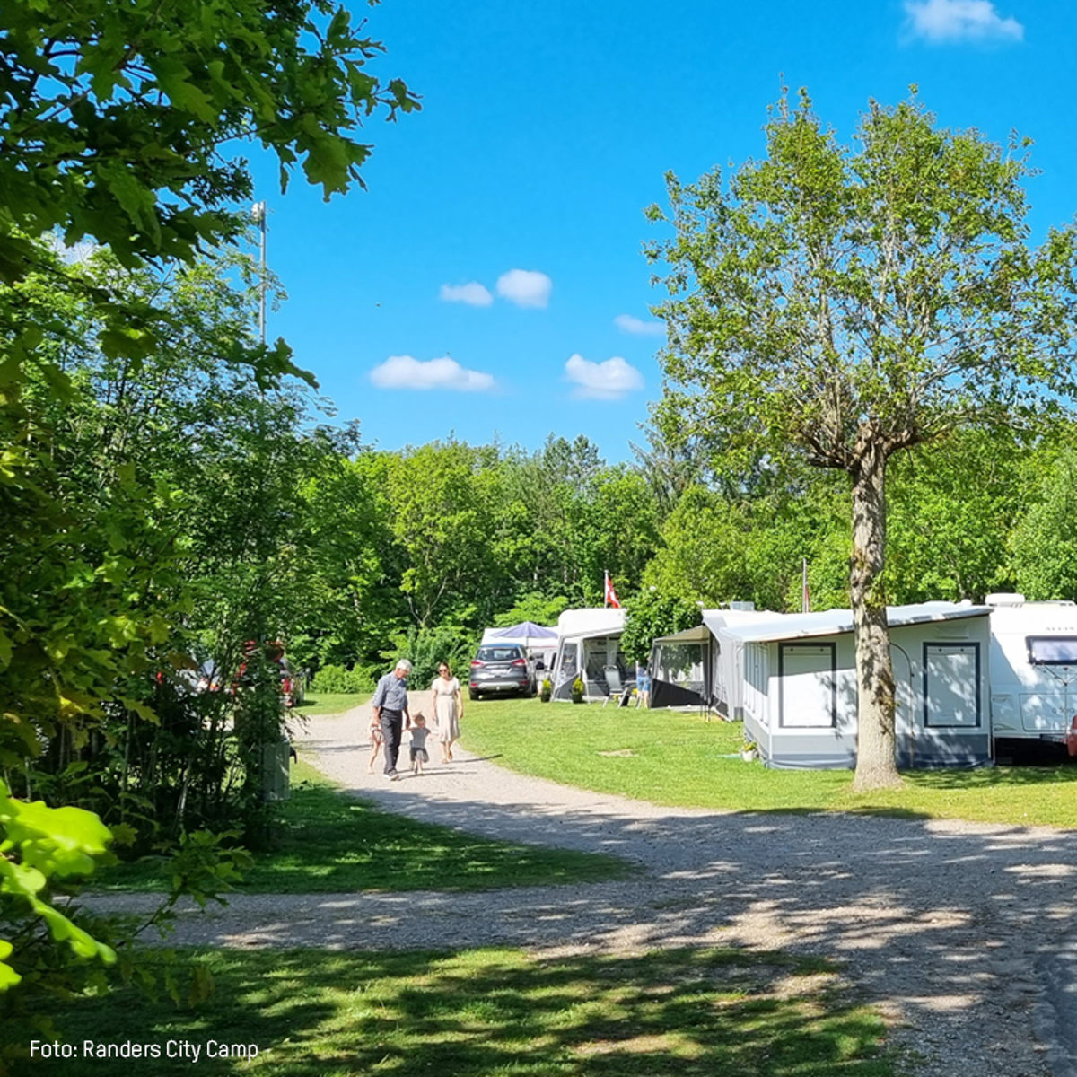 Familie går på grusvej gennem campingplads med campingvogne og telte omkranset af grønne træer og blå himmel.