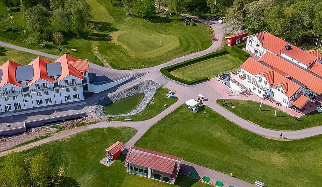 Golfbane og klubhus med røde tage i fugleperspektiv omgivet af grønne træer og velplejede plæner.