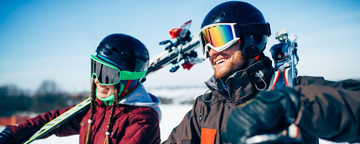 To personer med skibriller ses med store smil og sol i hovedet.