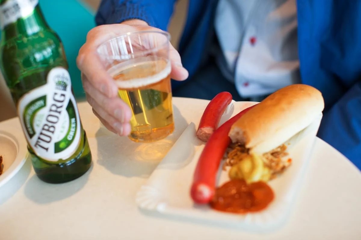 Mand holder plastikkop med lys øl ved siden af Tuborg flaske og rød pølse med brød og sennep på tallerken.