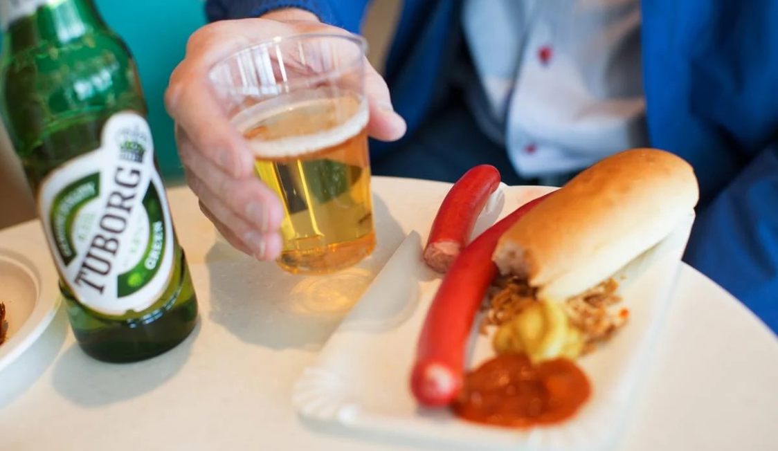 Mand holder plastikkop med lys øl ved siden af Tuborg flaske og rød pølse med brød og sennep på tallerken.