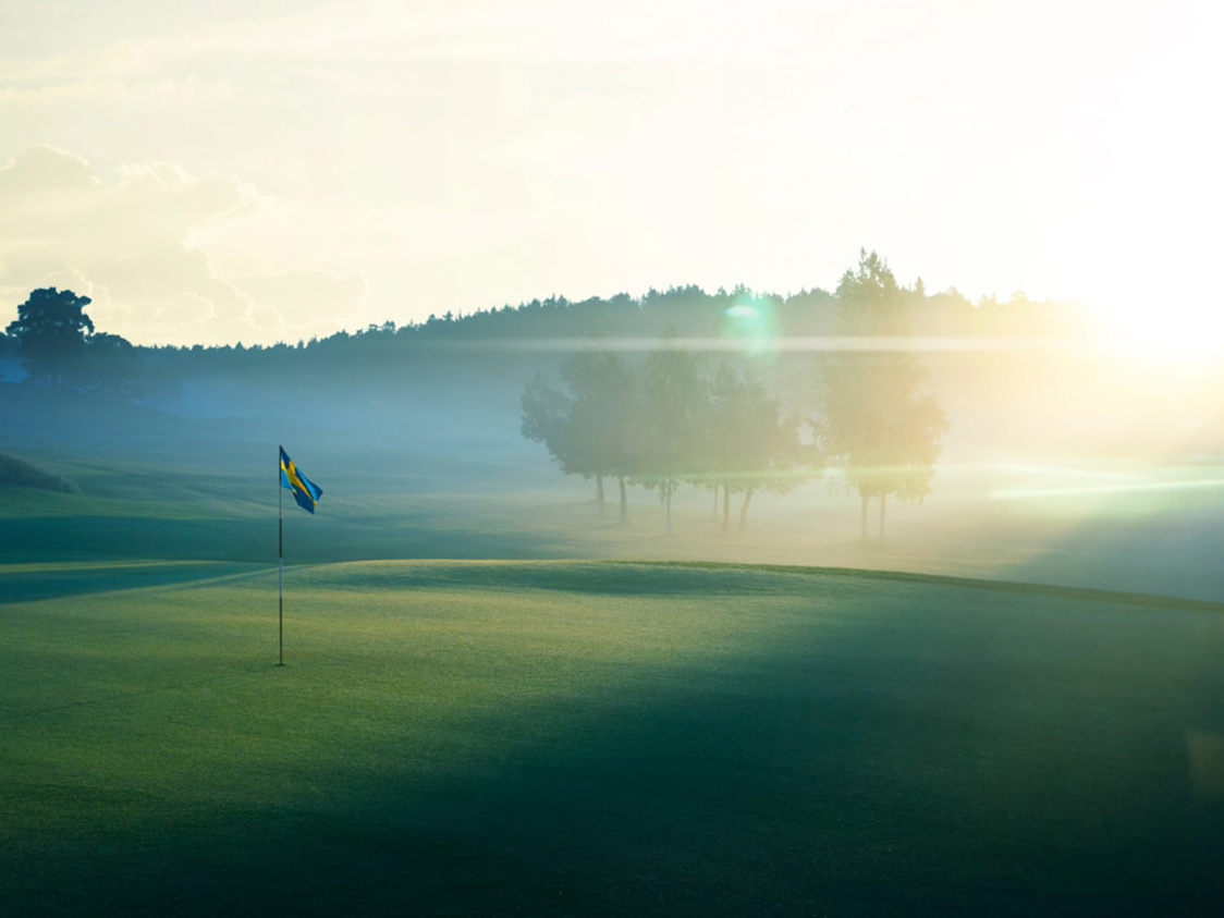 Svensk flag på en golfbane i solopgang med tåge omkring, set fra sidelinjen.