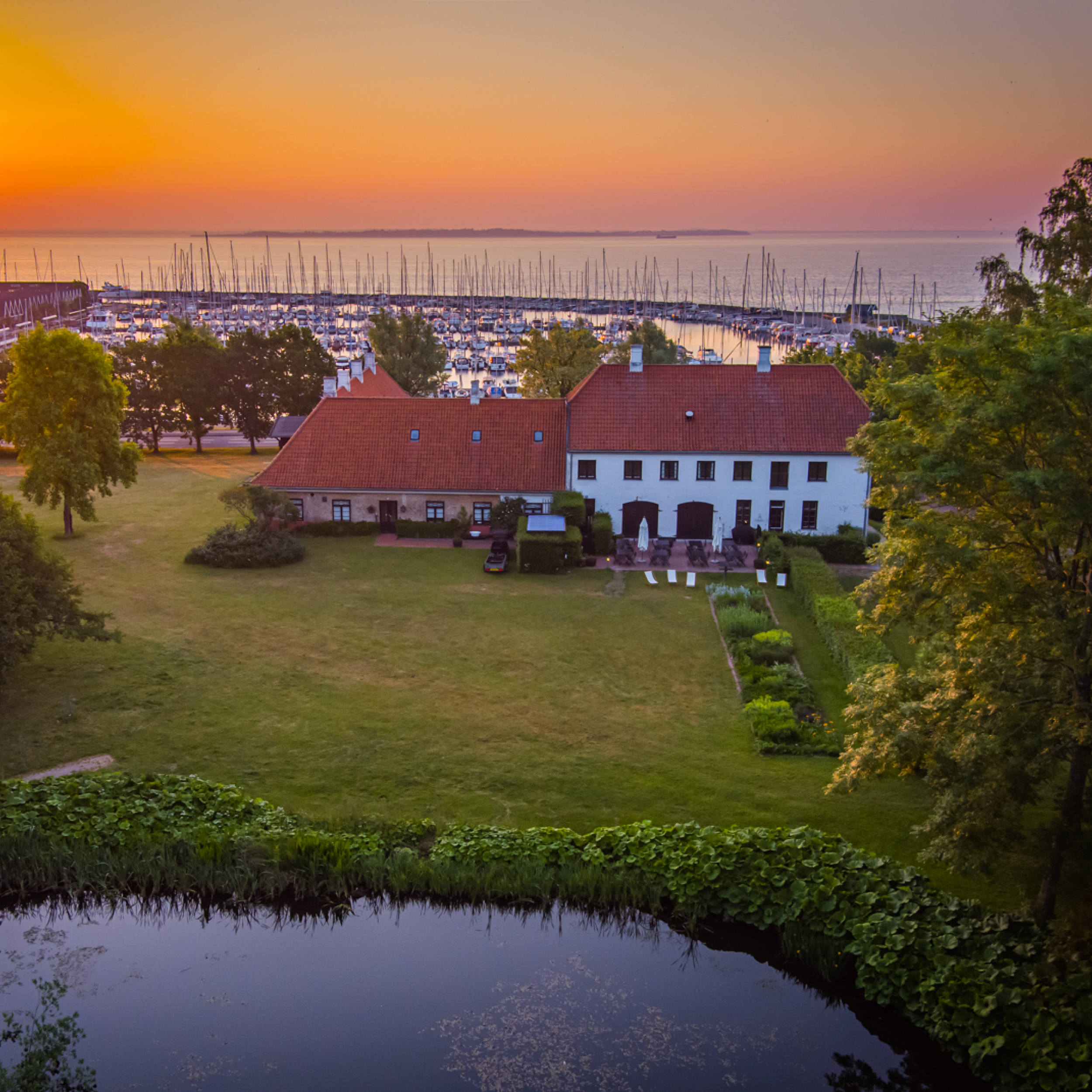 Historisk herregård ved smuk solnedgang, omgivet af grønne haver og med udsigt over en lystbådehavn.