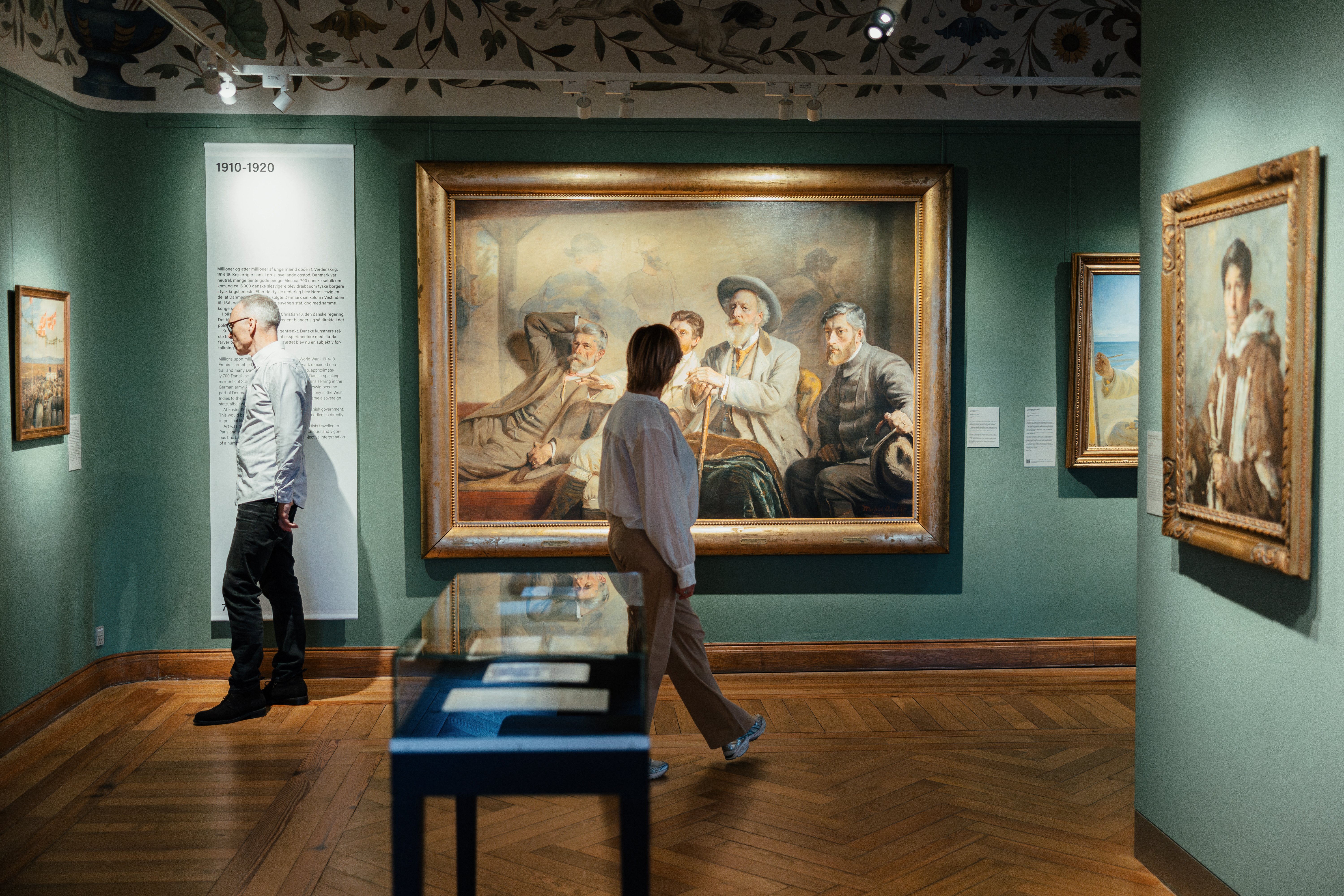 Besøgende på museum betragter stort maleri i guldindrammet ramme på udstillingsvæg.