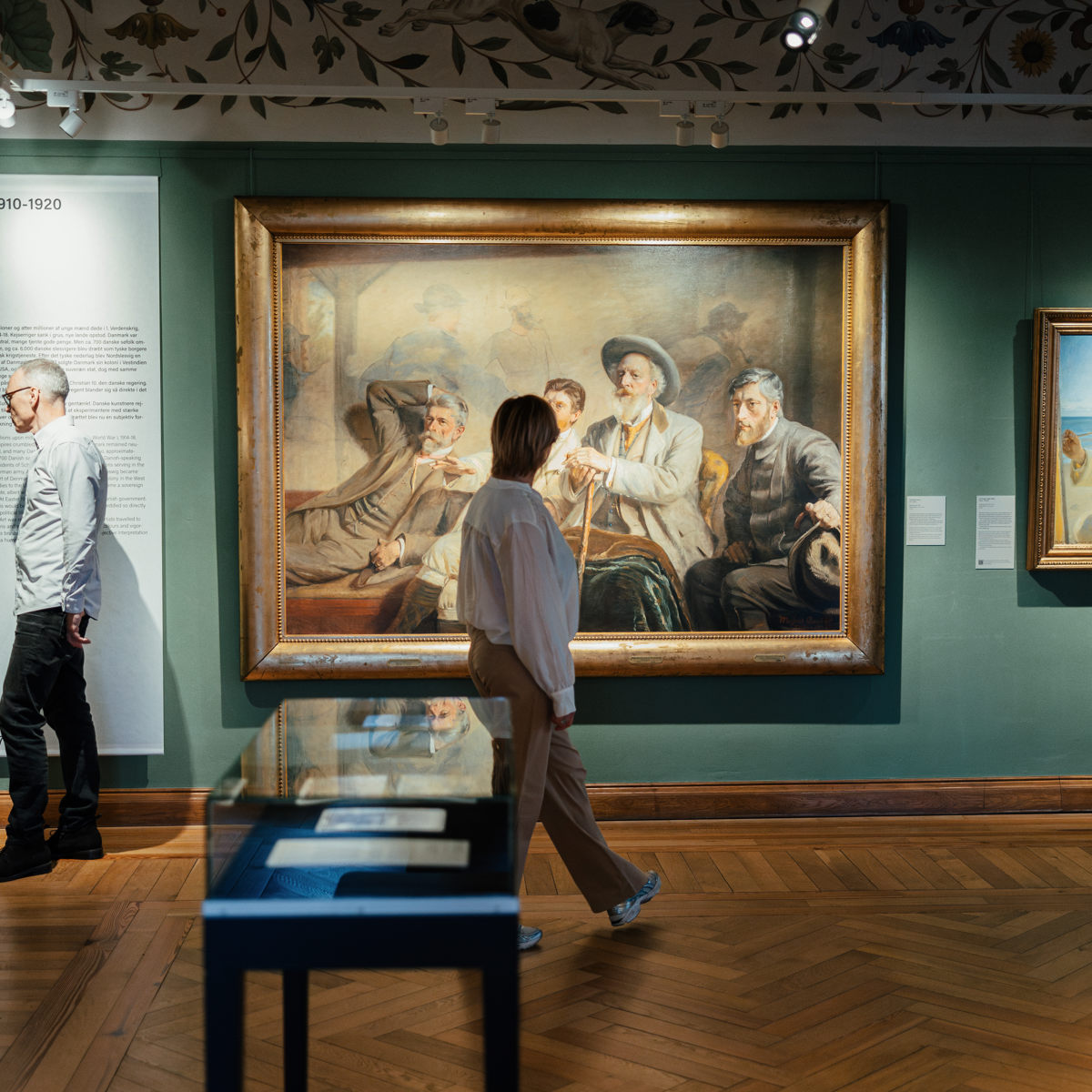 Besøgende på museum betragter stort maleri i guldindrammet ramme på udstillingsvæg.