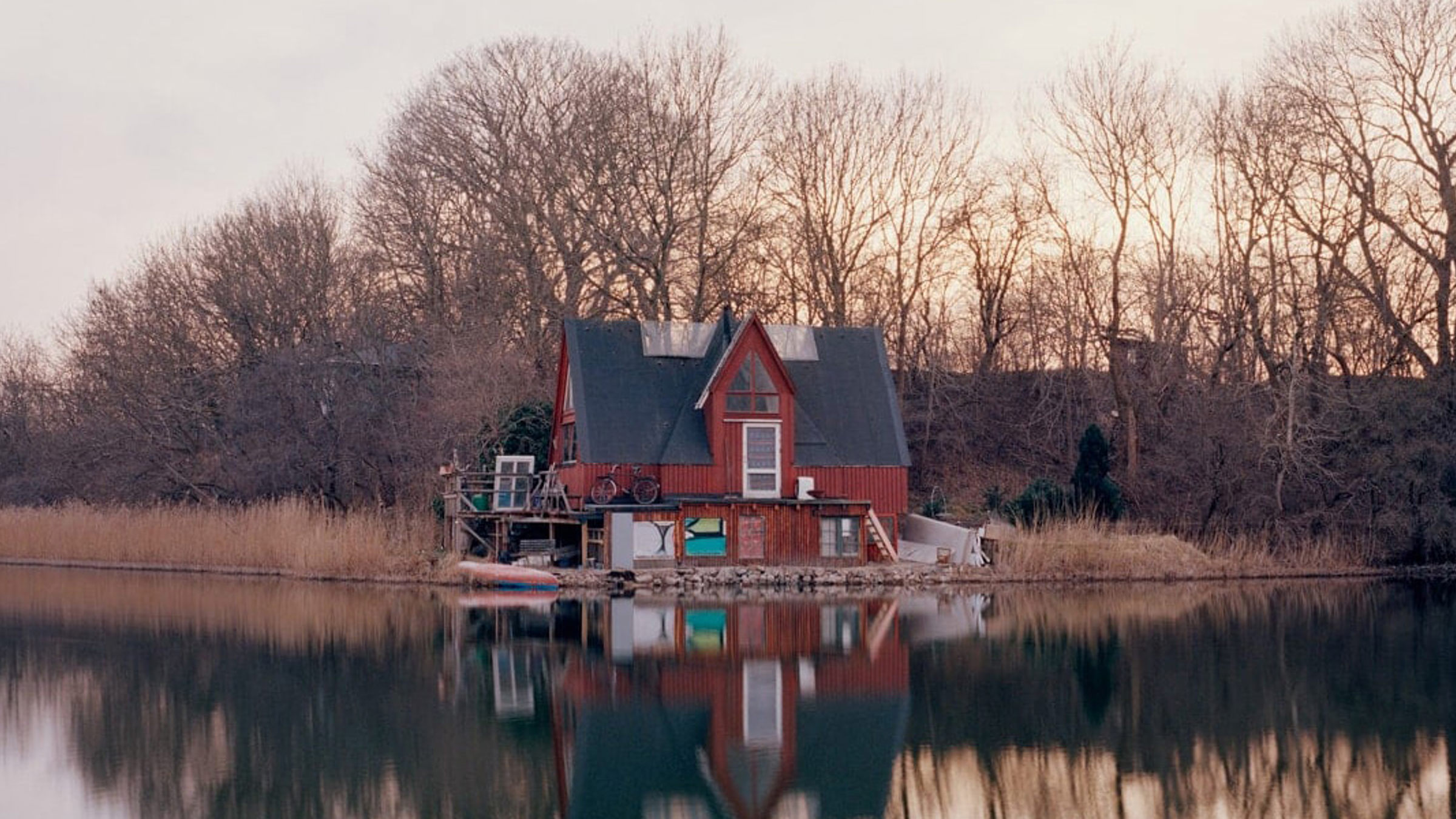 Rött hus vid vatten med spegelbild i sjön och skog i bakgrunden, under en stilla solnedgång.