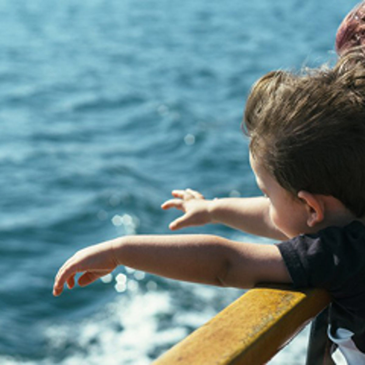 Dreng kigger ud over skibsgelænder mod havet med udstrakte arme under en solrig dag.
