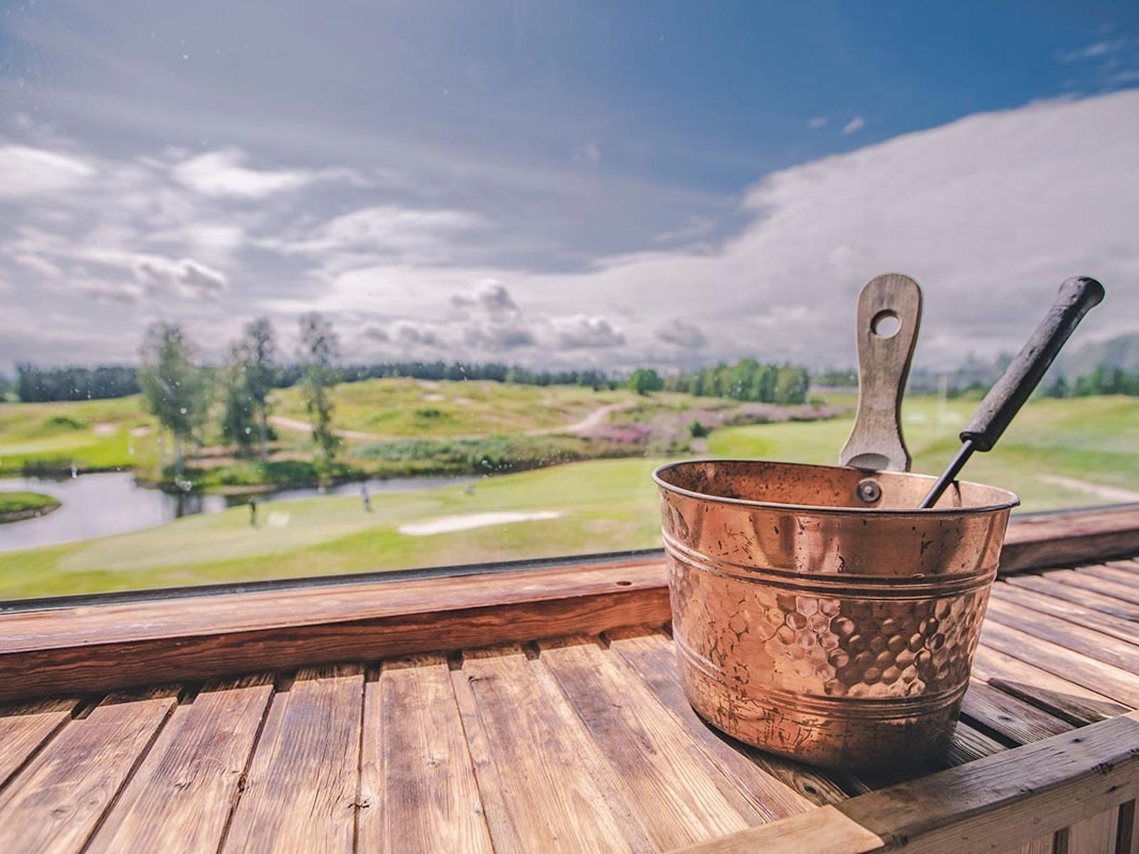Kobberspand med metaltang står på træbord foran udsigt til golfbane og skov.