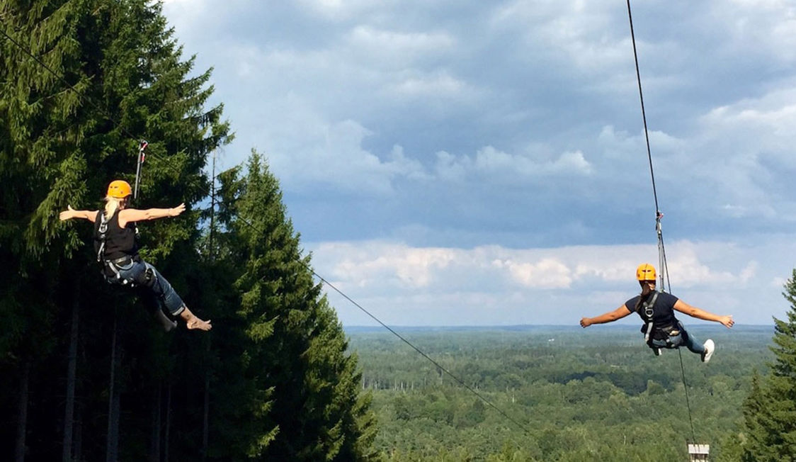 To personer iført sikkerhedsudstyr svæver på en svævebane over en skov med grønne træer i baggrunden.
