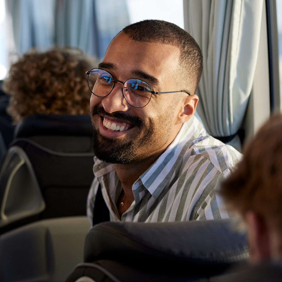 En man på en buss ser glad ut iförd randig skjorta och glasögon, i en ljus och bekväm sittplats.