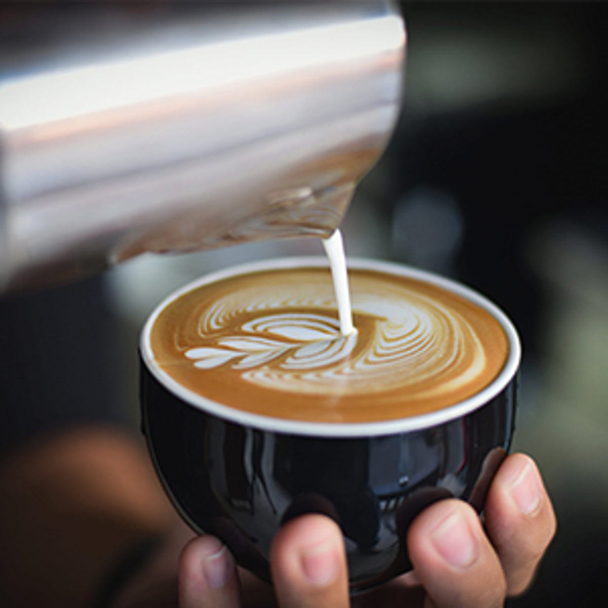 Barista hælder skummet mælk i cappuccino med latte art mønster i en sort kop, set fra siden.