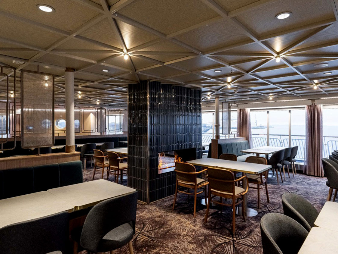 Moderne færgesalon med stilfulde træmøbler, lys i loftet og store vinduer der viser havudsigt.