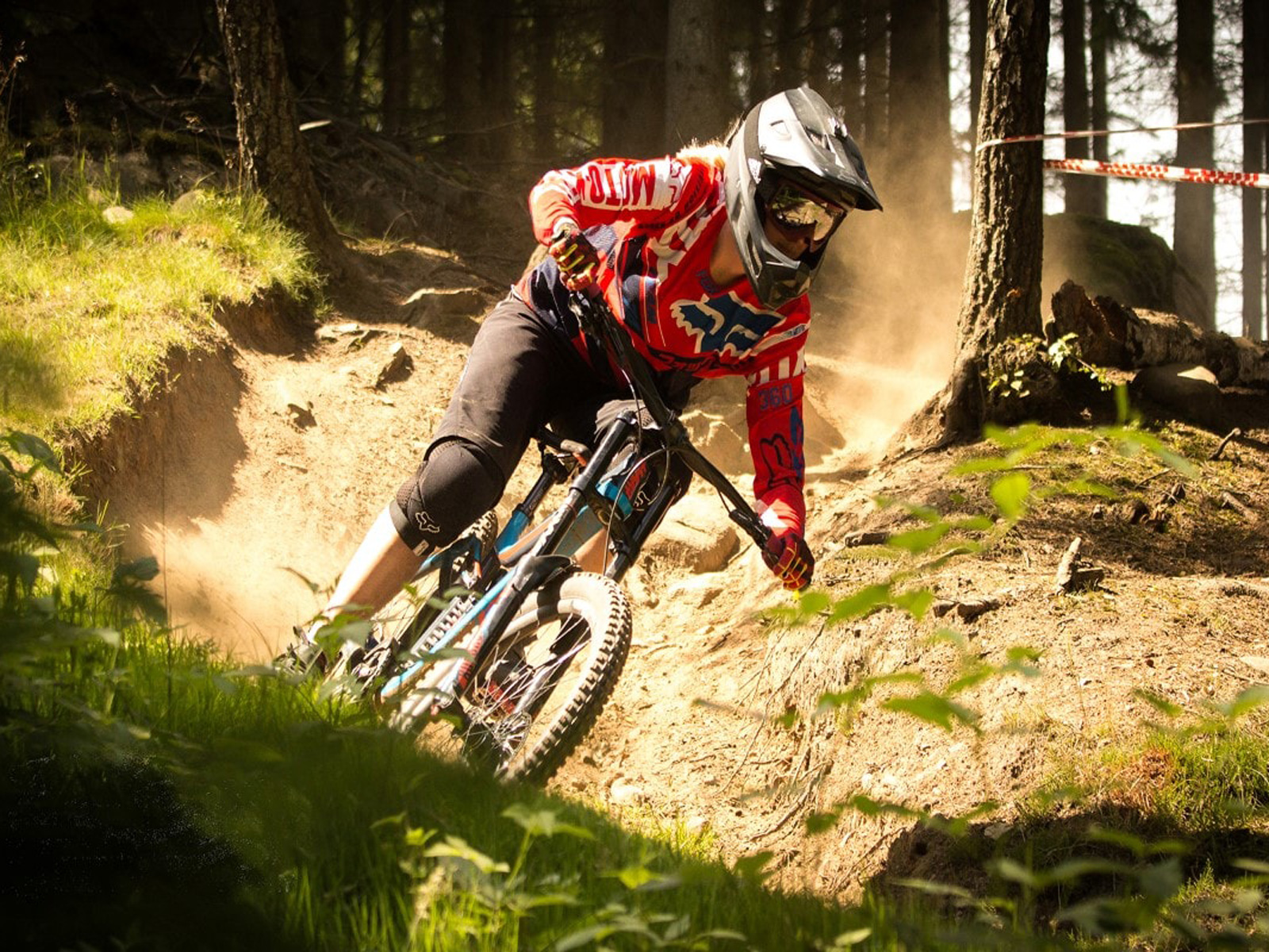 Mountainbike-rytter med rød trøje kører ned ad en skovsti med skrånende kurver og sollys gennem trækronerne.