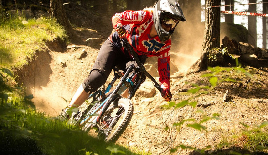 Mountainbike-rytter med rød trøje kører ned ad en skovsti med skrånende kurver og sollys gennem trækronerne.