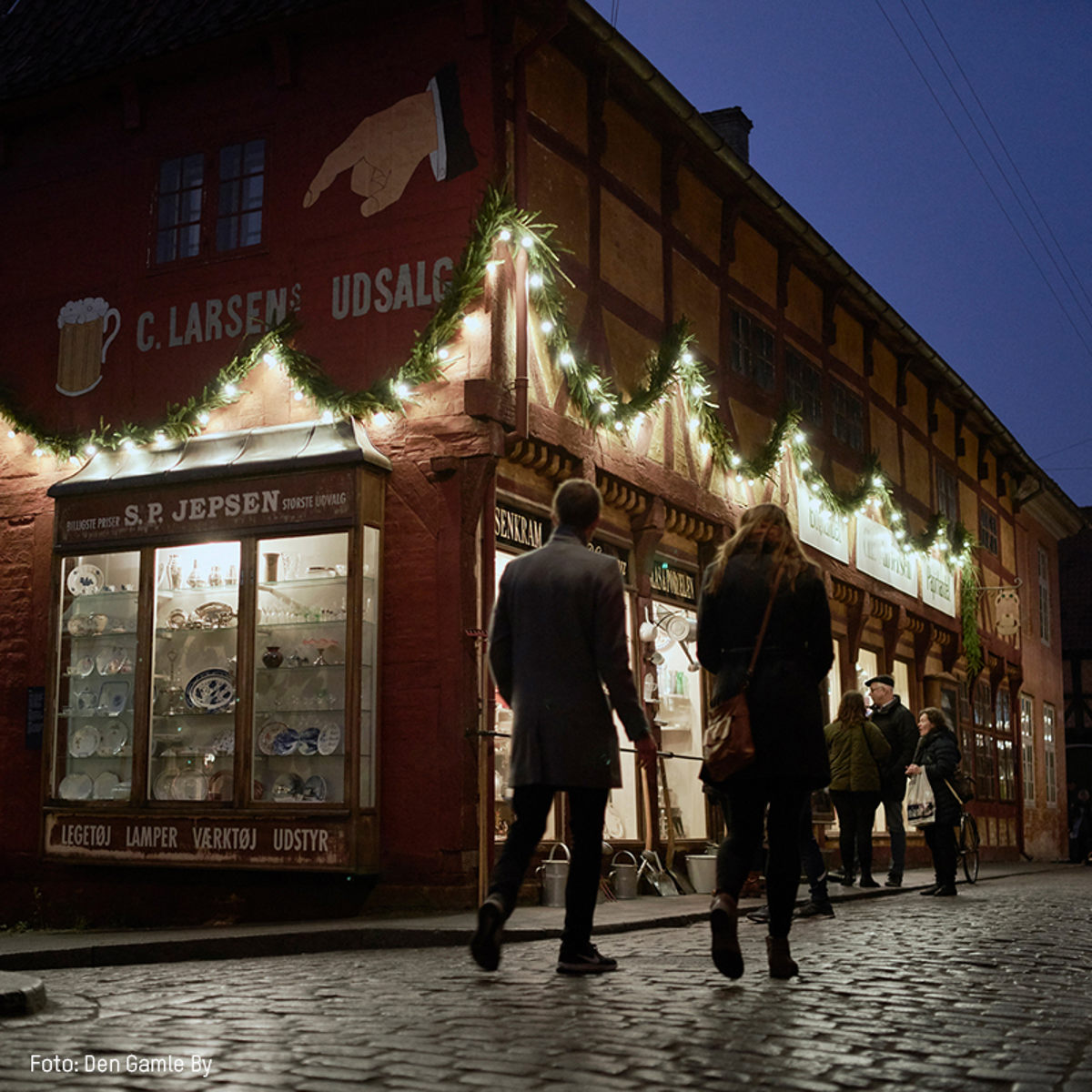 Personer går forbi en oplyst butik i en gammel by, dekoreret med julelys og granguirlander om aftenen.