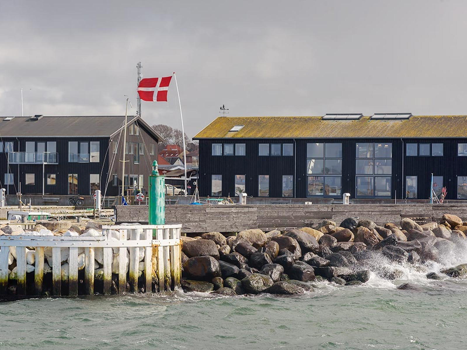 Dansk flagga vajar vid Hundested hamn med vågor som slår mot stenarna i en kustnärbild.