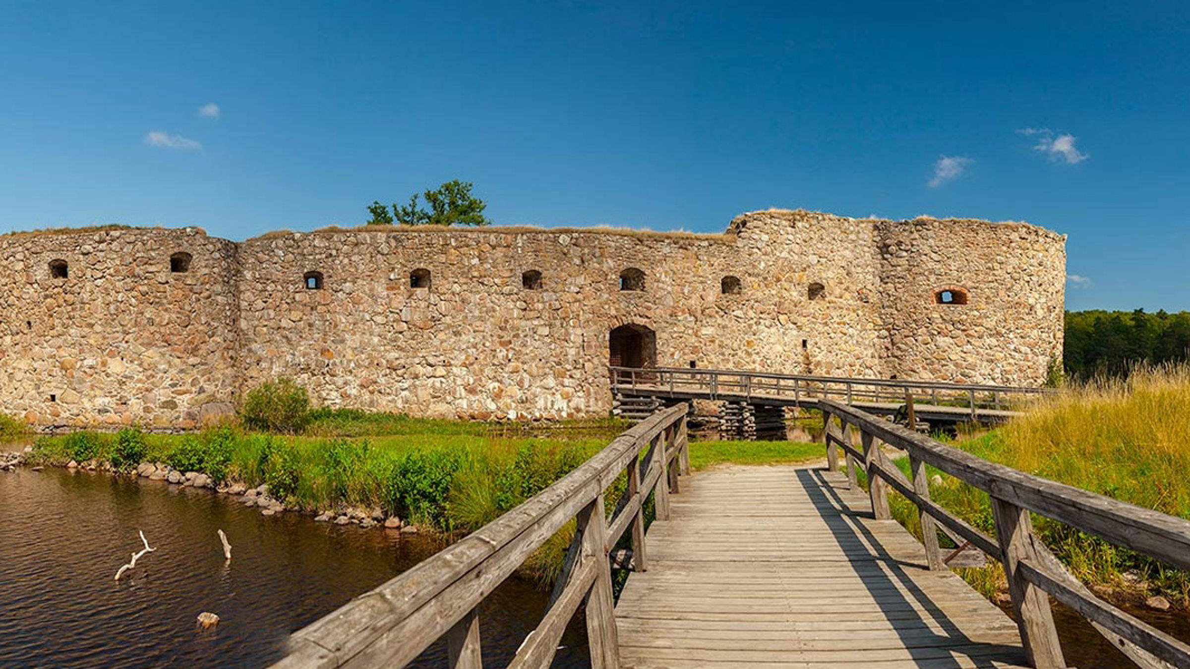 Ruinen af middelalderborg omgivet af grønt landskab og blå himmel set fra træbro.
