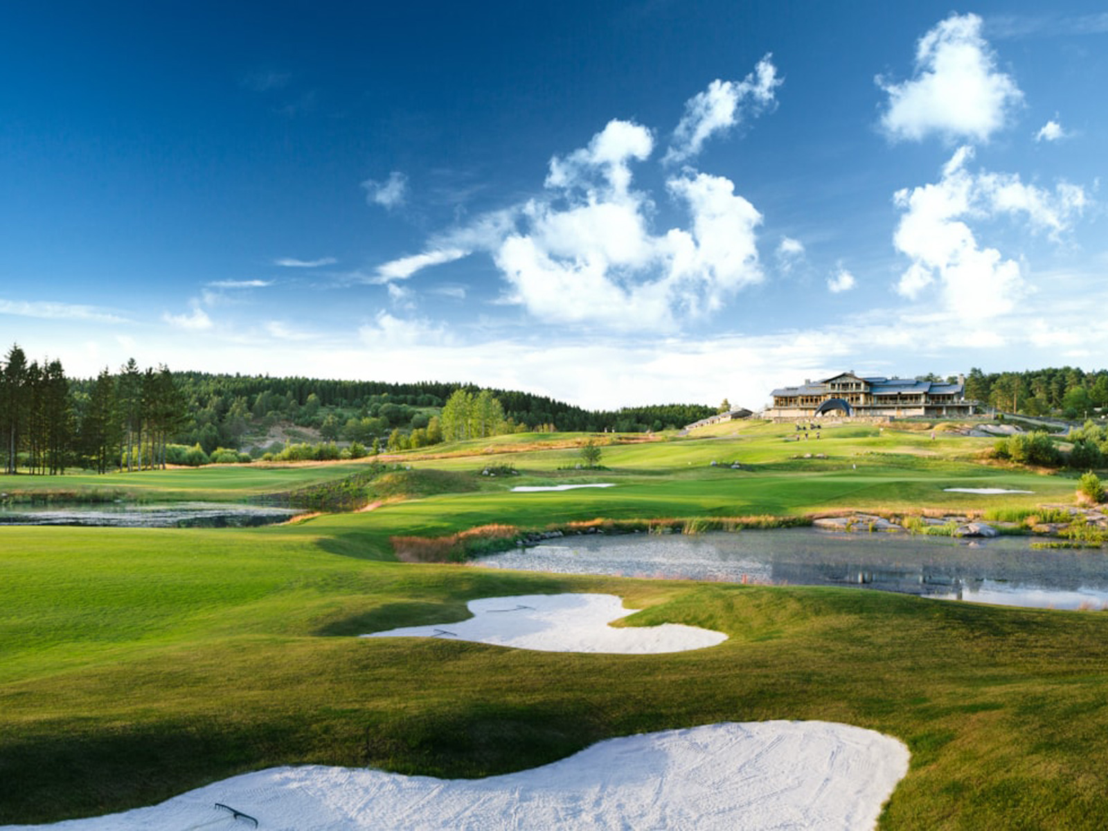 Golfbane med grønne fairways og klar blå himmel i fugleperspektiv, klubhus i baggrunden.