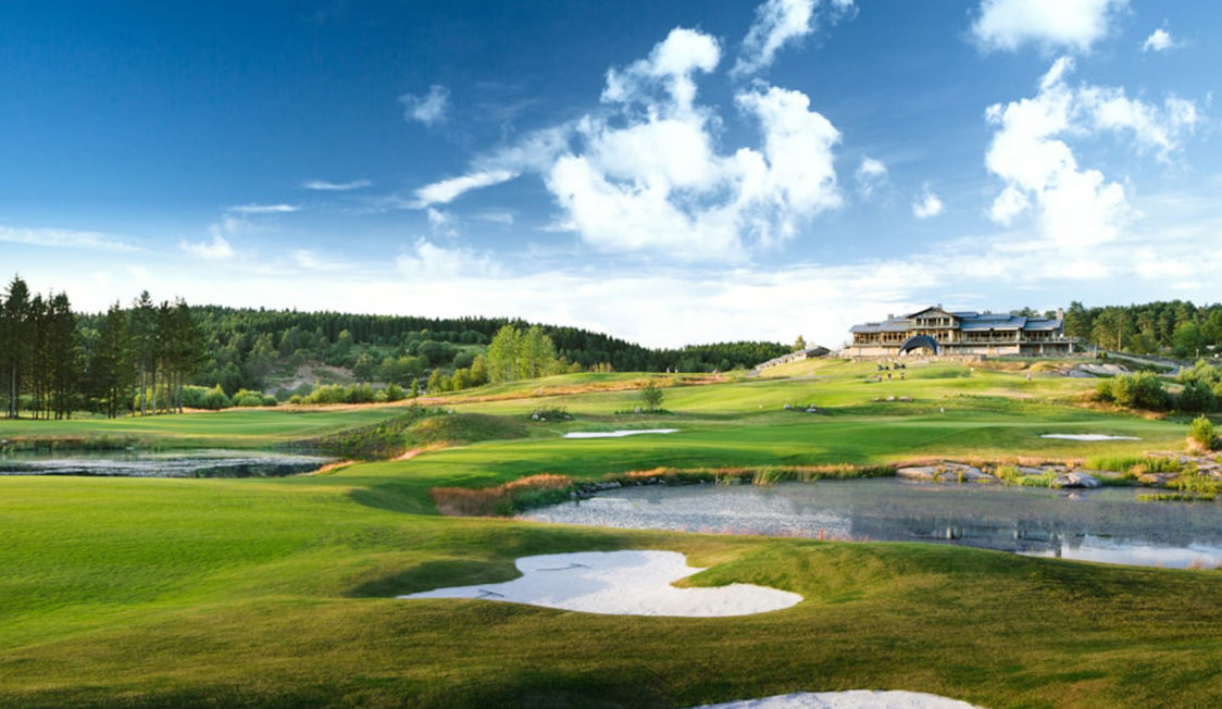 Golfbane med grønne fairways og klar blå himmel i fugleperspektiv, klubhus i baggrunden.