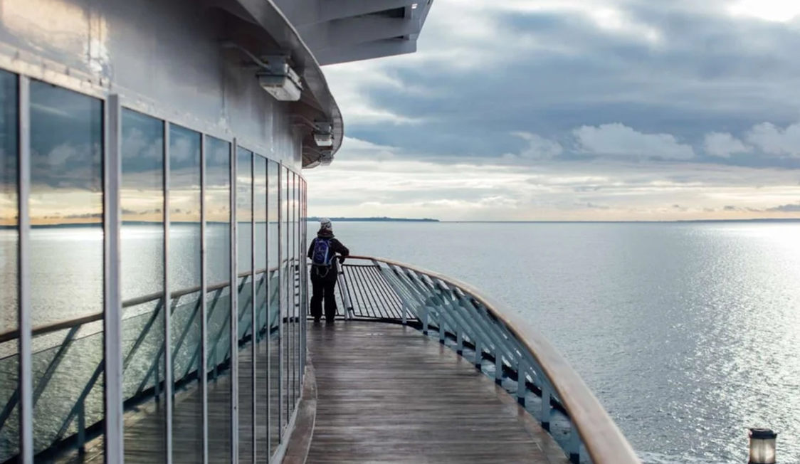 Person står ved rælingen på et skib, med havudsigt og reflekteret lys på panoramaglasset.