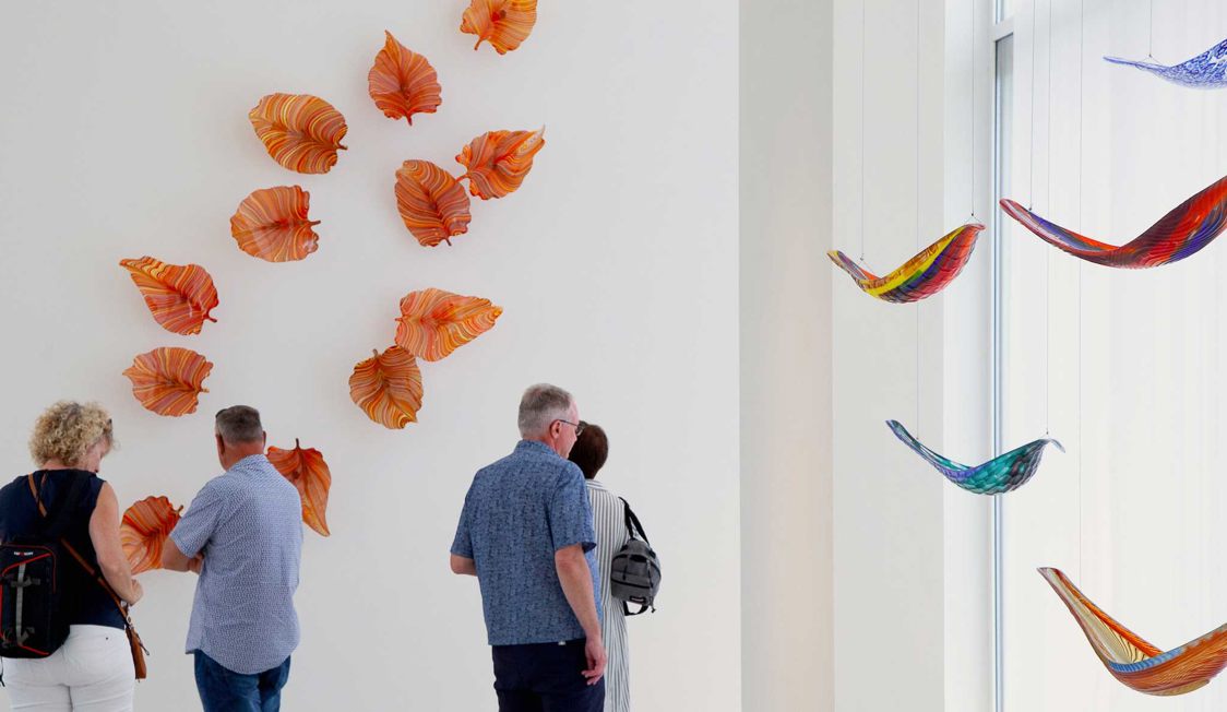 Værk af orange glasblade på væggen i et kunstgalleri hvor folk betragter kunstværket.