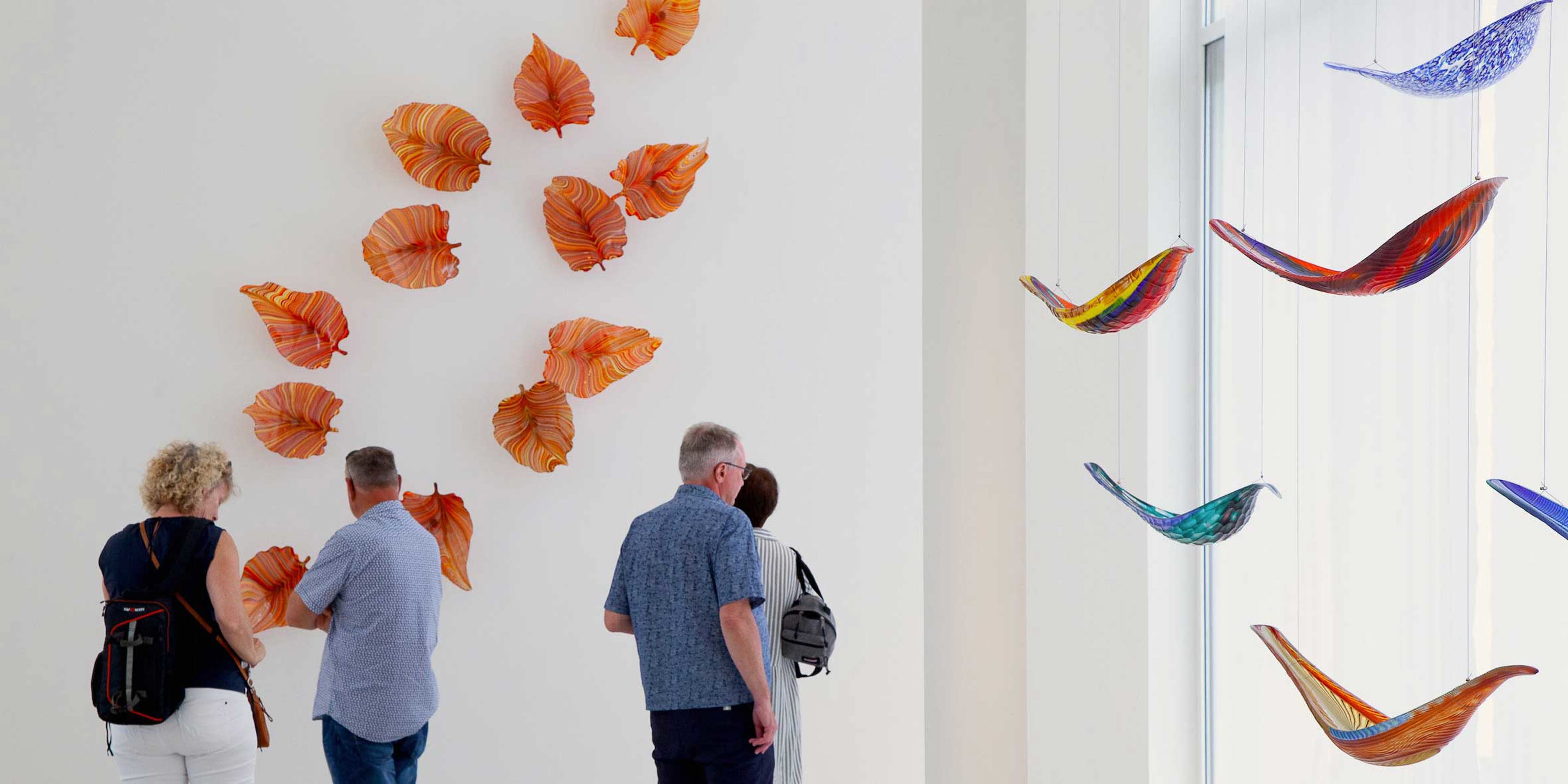 Artwork of orange glass leaves on a gallery wall with people observing the installation.