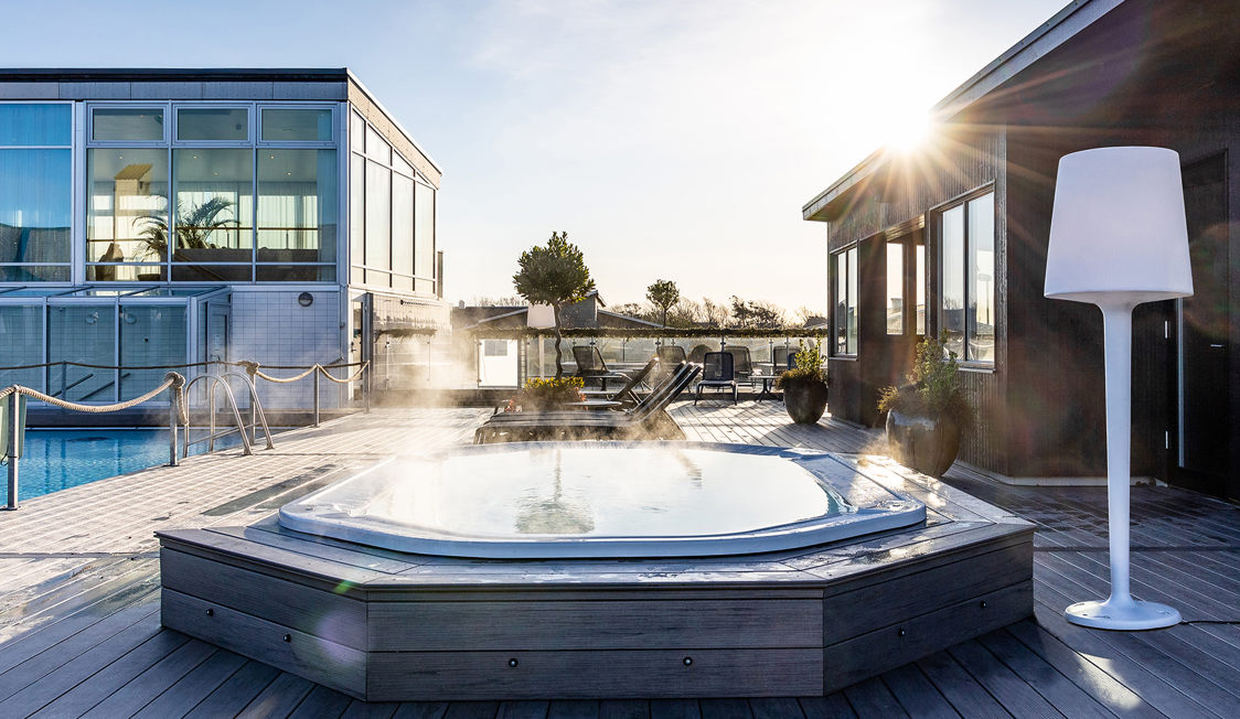 Udendørs jacuzzi med dampende vand på træterrasse i solskin nær moderne bygning.