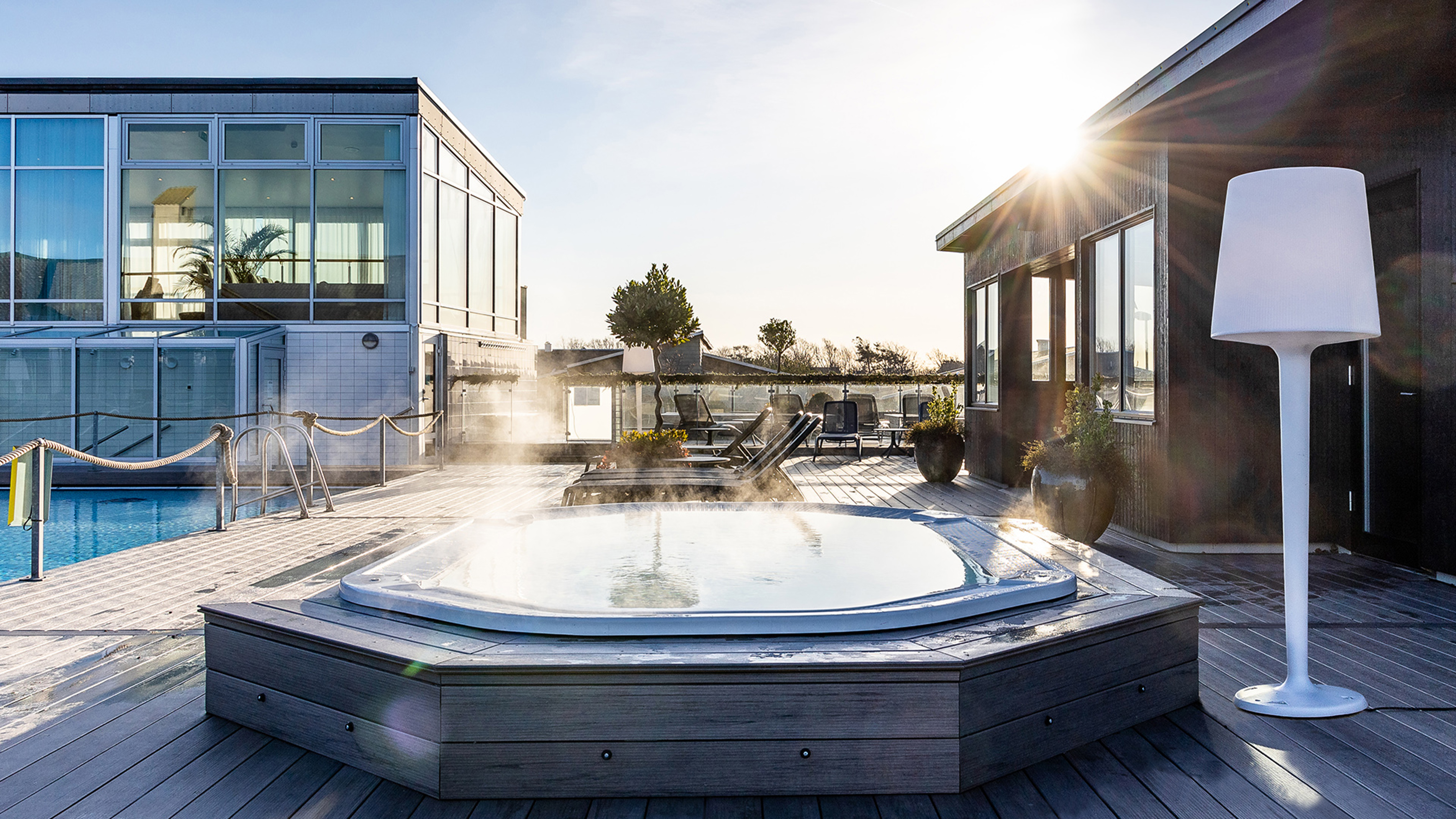 Udendørs jacuzzi med dampende vand på træterrasse i solskin nær moderne bygning.