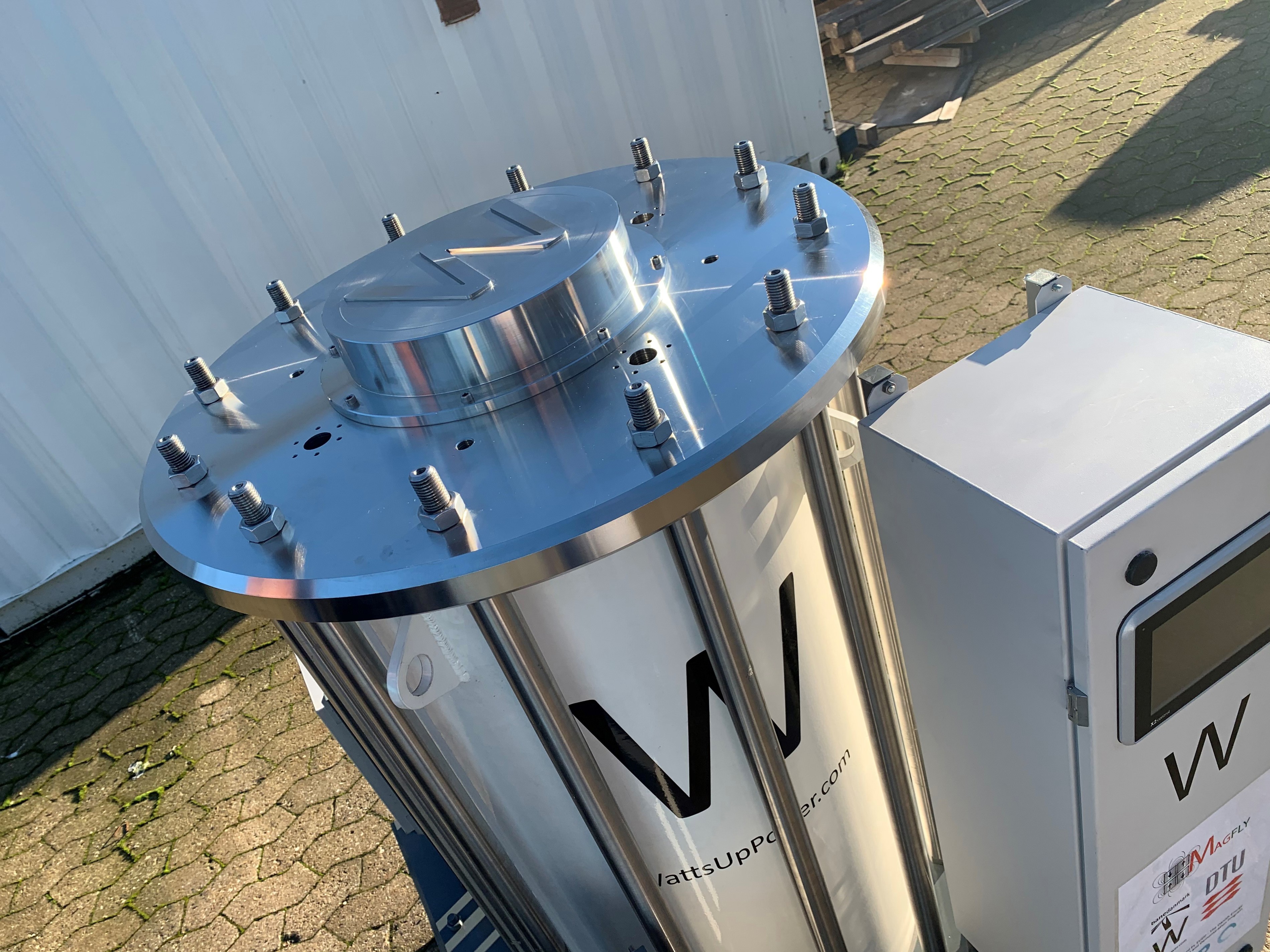 Eksperimentelt sølvmaskineri med logo for WattsUpPower står udendørs på fortovet ved siden af en kontrolboks.