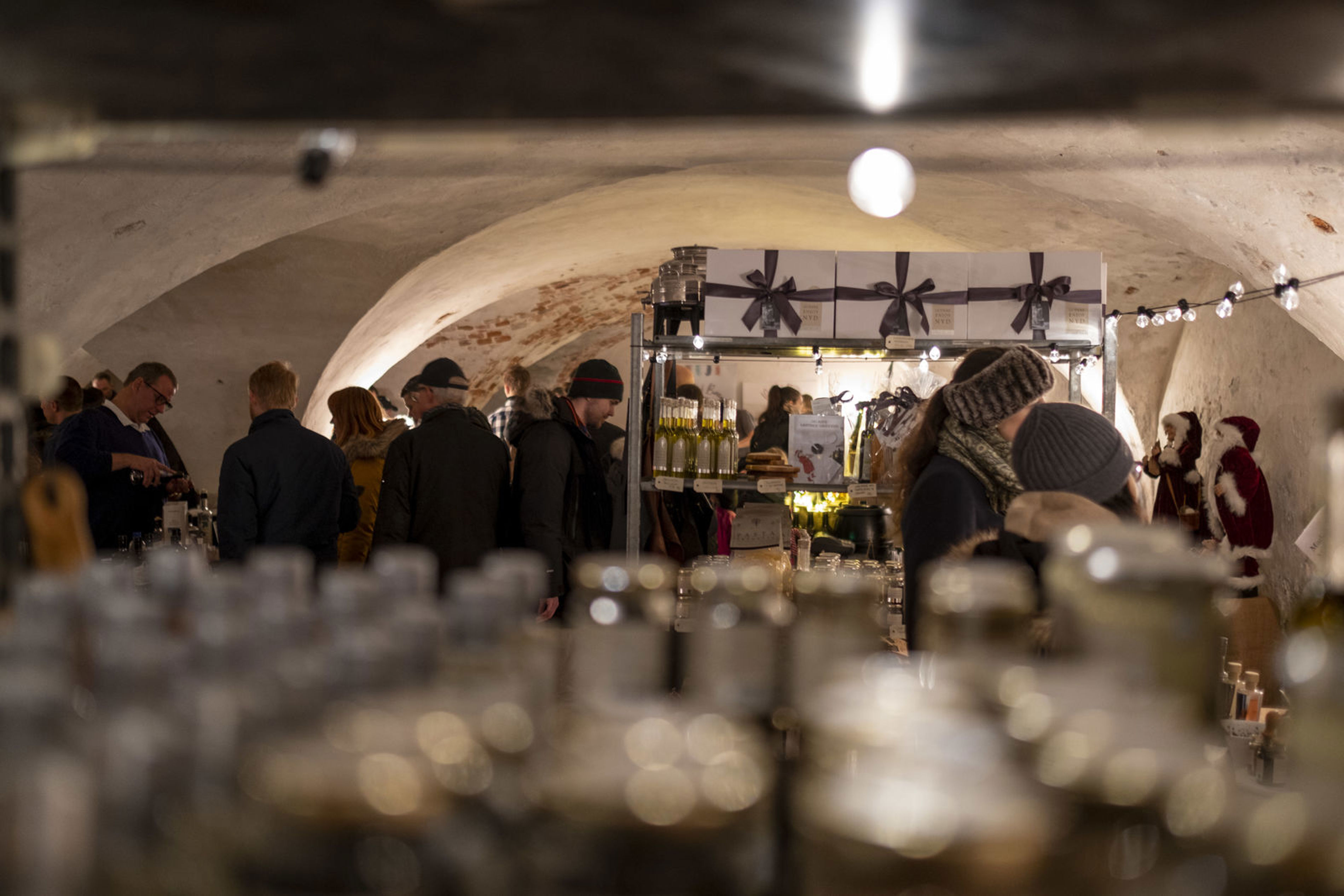Folk besøger stand med vine og delikatesser i en hyggelig kælder, oplyst af varm belysning under marked.
