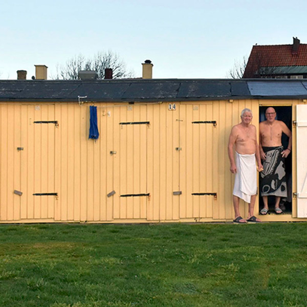 Ældre mænd ved åbning af gul træbadehus på grøn græsplæne, iført badehåndklæder.