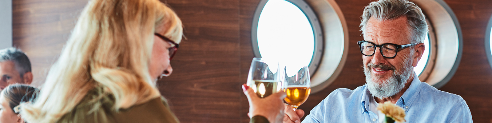 Par skåler i glas med vitvin, leende mot varandra på en färja, bruna träpaneler och runda fönster i bakgrunden.