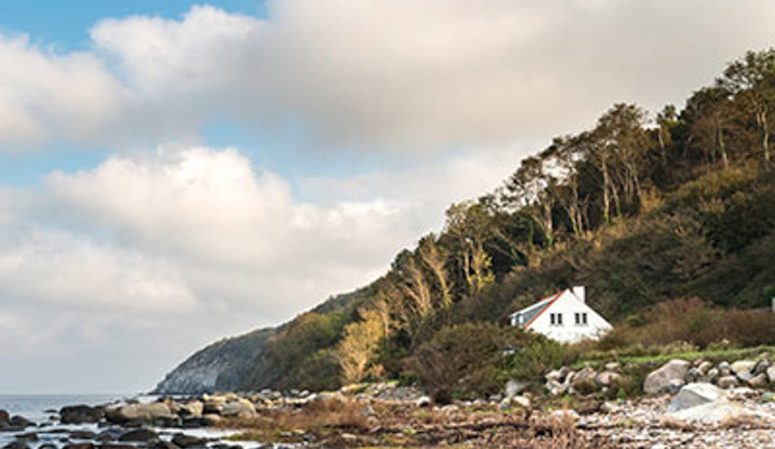 Hvidt hus beliggende ved skovklædt kystlinje med havudsigt på Bornholm under en overskyet himmel.